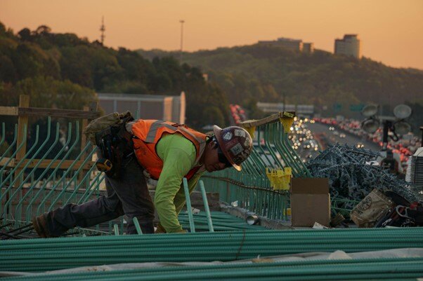 Construction worker in a hard hat and orange vest working on rebar installation at sunset, with a busy highway and cityscape in the background.}