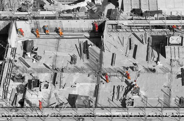 Overhead view of construction workers in orange uniforms working on a large construction site with various equipment and materials.