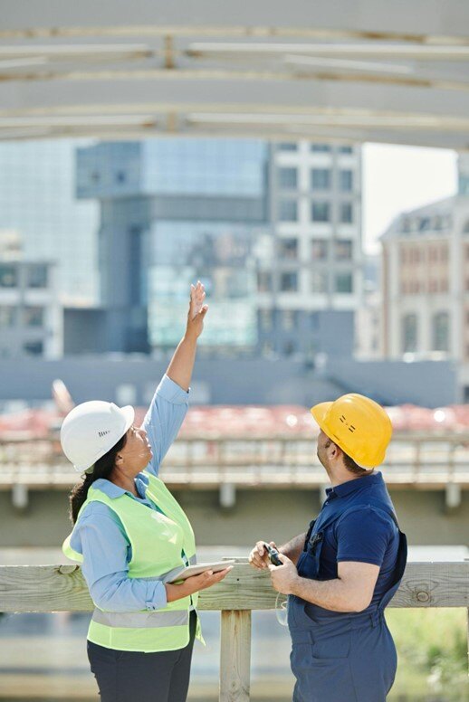 Two construction workers wearing safety gear discuss a building project outdoors, with one pointing upwards and the other holding a tool